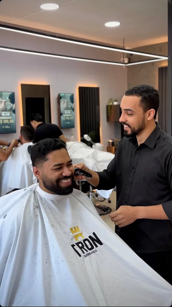 Barbeiro sorridente finalizando o corte com máquina enquanto o cliente, vestindo a capa TRON, sorri satisfeito